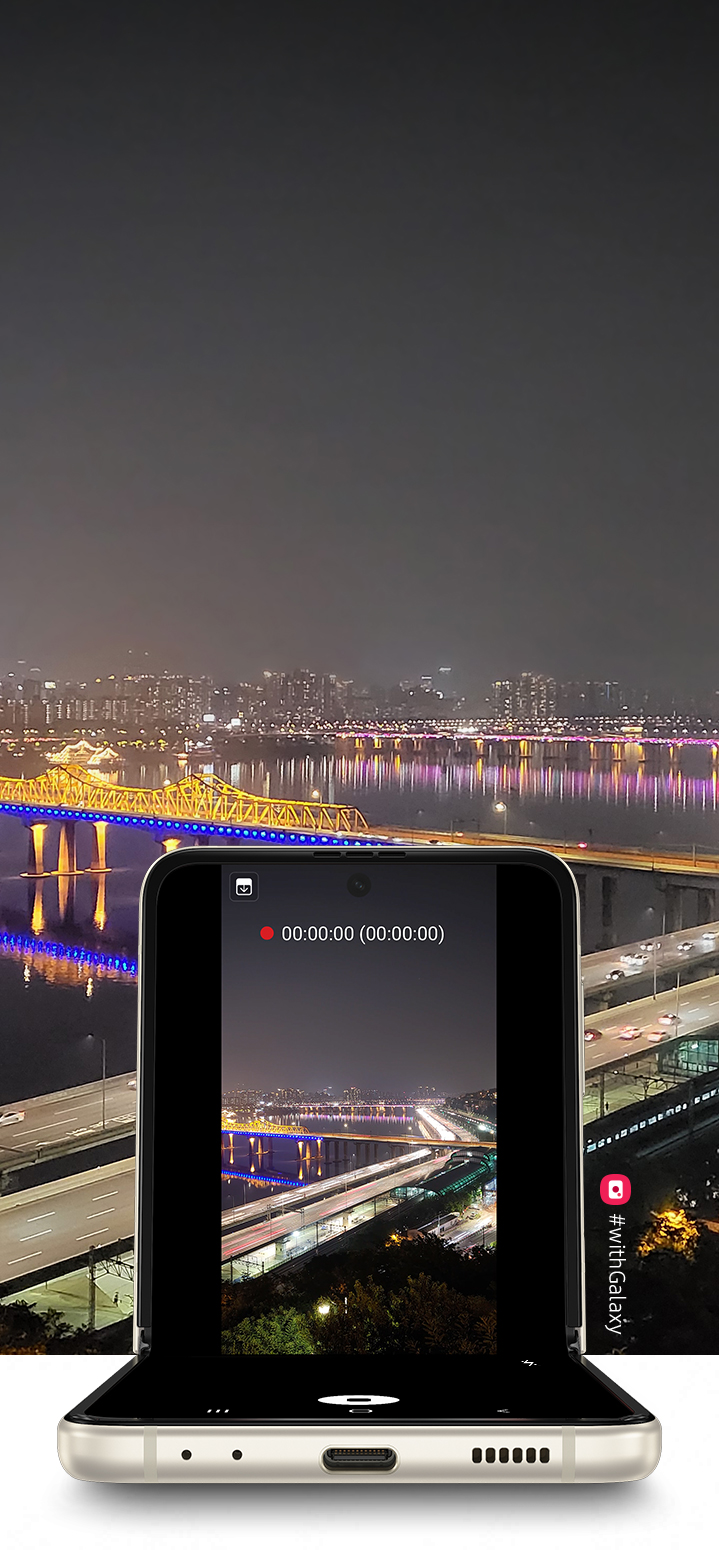 A view of a cityscape at night, as seen from the top of a hill. Galaxy Z Flip3 5G in Flex mode with the Camera app on the Main Screen and the same cityscape in the viewfinder, being recorded with Night Hyperlapse.