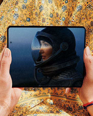 Hands holding Galaxy Z Fold3 5G, unfolded. The Main Screen displays a scene from a game of a woman in a space helmet.