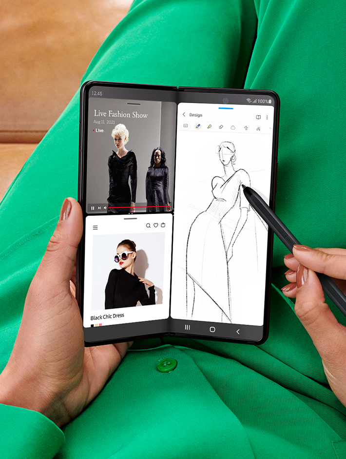 Hands holding unfolded Galaxy Z Fold3 5G and sketching with the S Pen Fold Edition. The Main Screen is in Multi Active Window mode. One half of the screen shows two instances of the Internet app open, one with a live stream of a fashion show and the other with a website showing images of the garments from the show. The other half of the screen is a sketch of a dress being done in Samsung Notes.